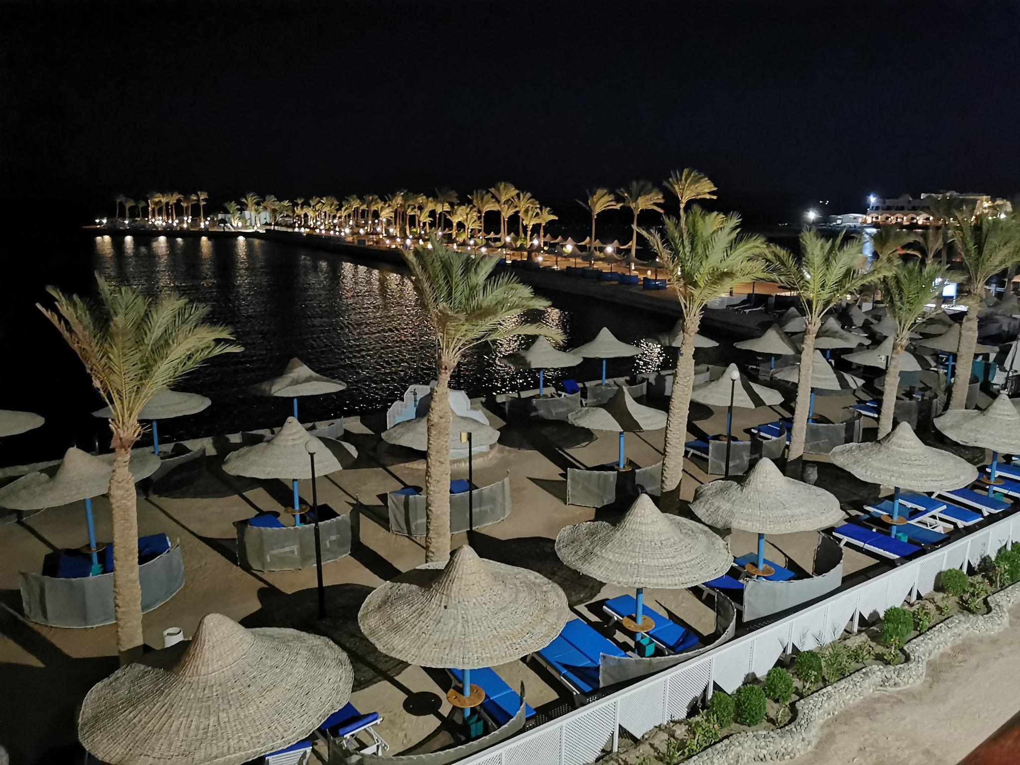 Illuminated beach resort at night with straw umbrellas, blue loungers, and palm-lined bay.