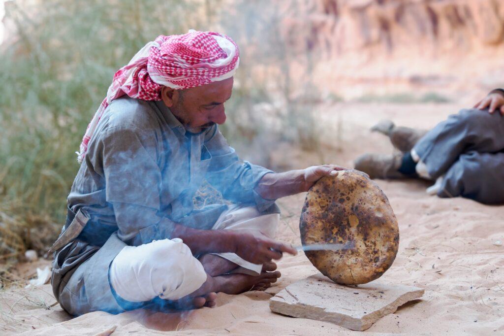 A Bedouin man in traditional keffiyeh baking flatbread over hot coals in the desert