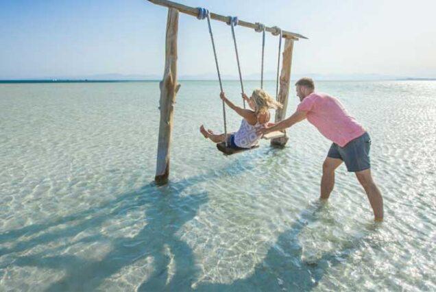 A man pushes a woman on a wooden swing set in shallow crystal-clear Red Sea waters