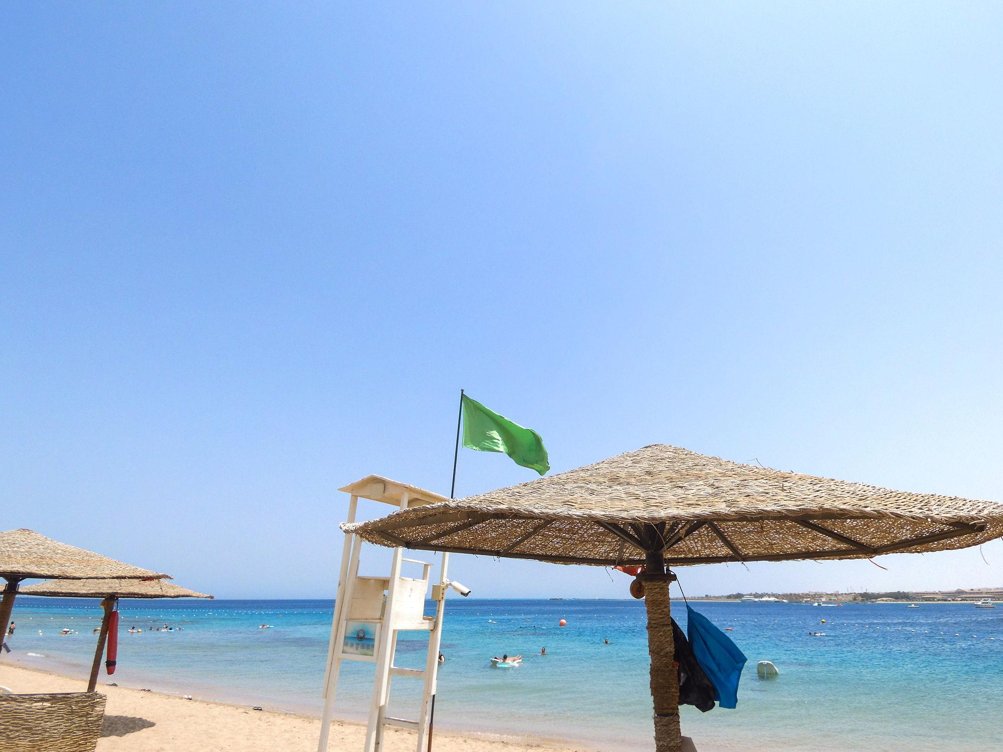 A green beach safety flag flies above a lifeguard tower beside thatched umbrellas on the Red Sea shore.