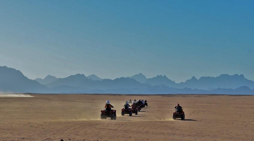 A convoy of quad bikes crossing a vast desert plain with mountains in the background