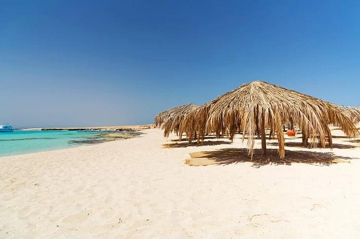 Row of thatched palm umbrellas lining a white sandy Red Sea beach under clear blue sky
