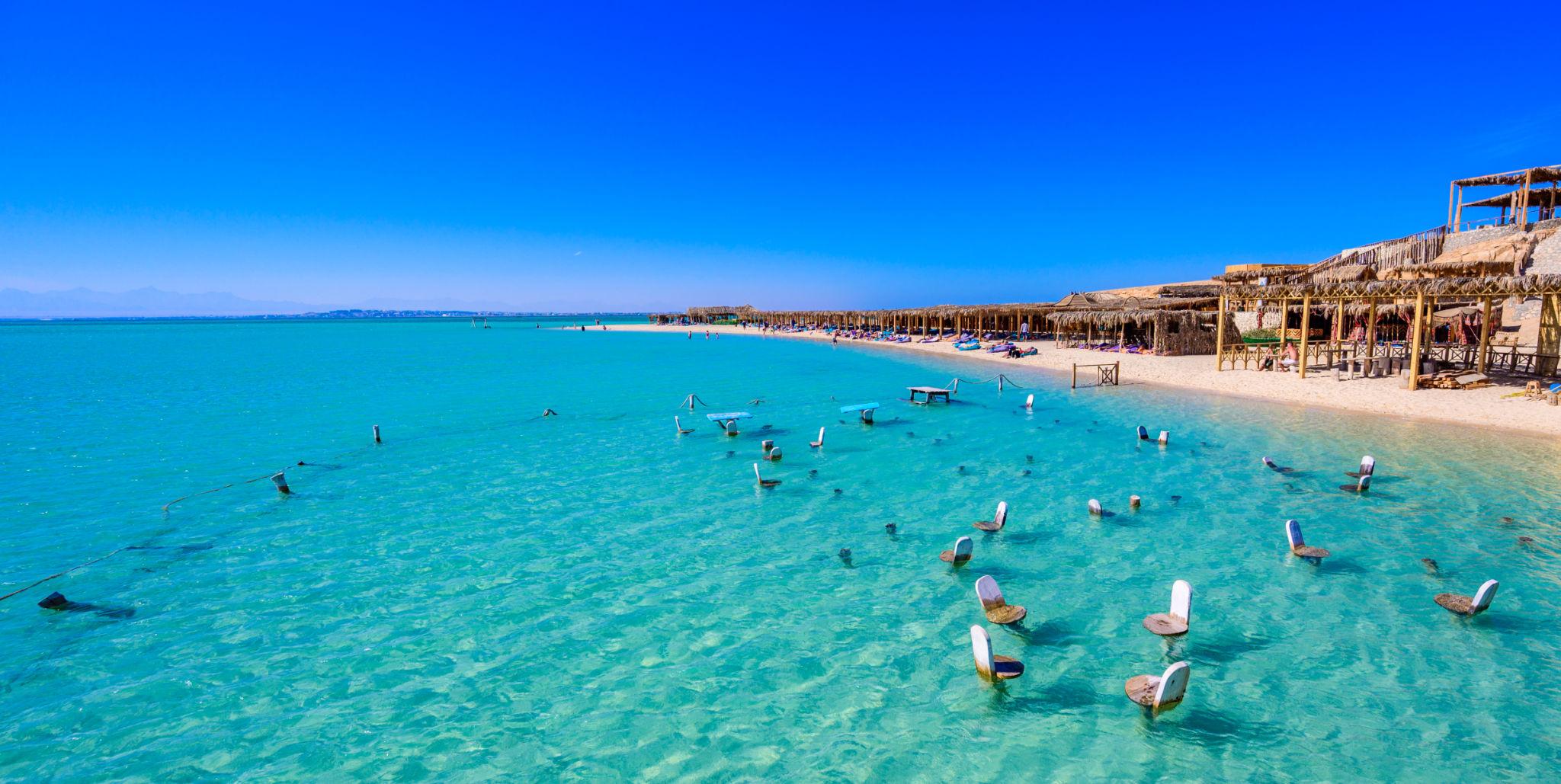 Submerged chairs dot a crystal-clear turquoise lagoon beside a thatched beach resort.