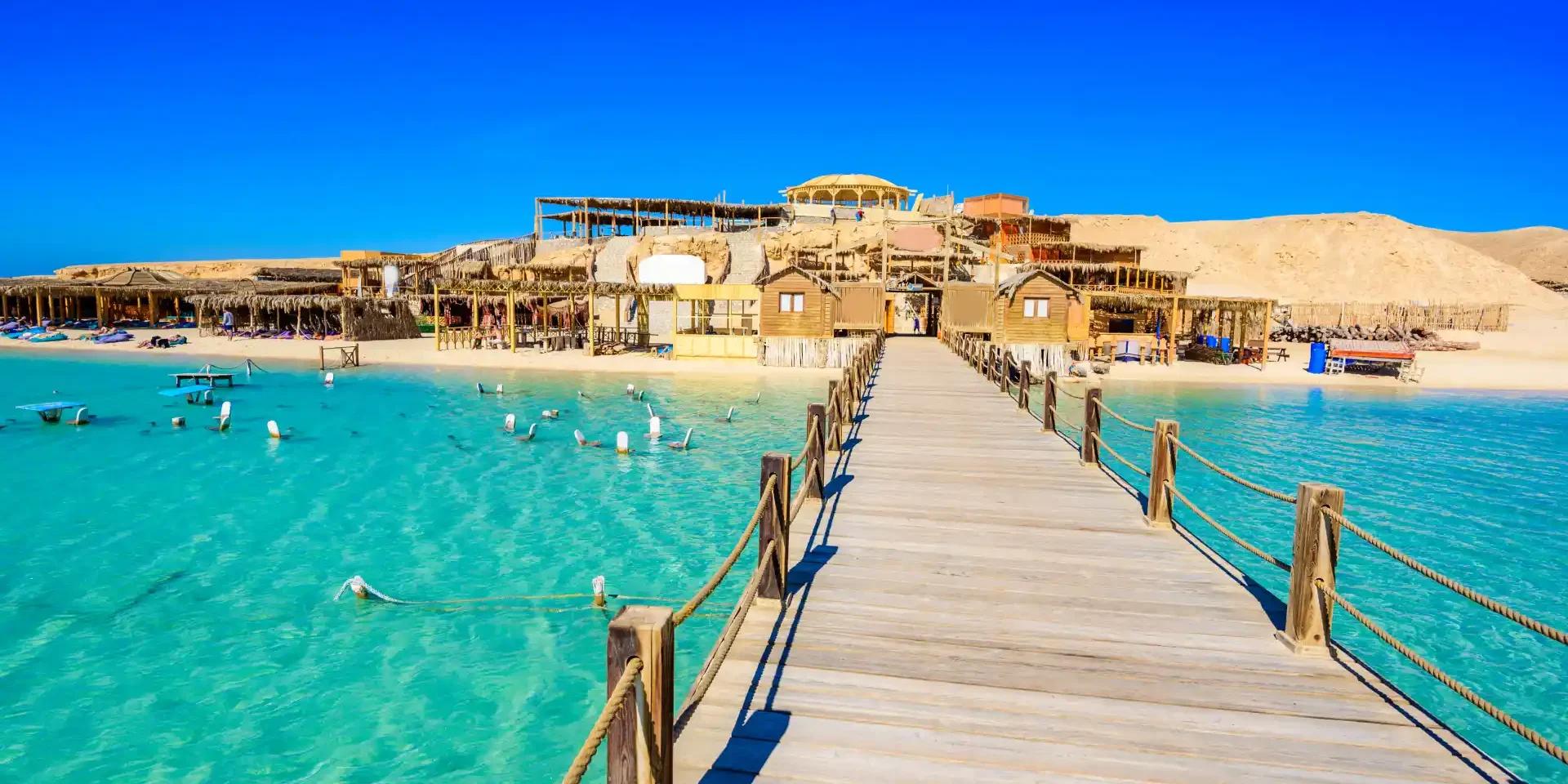 A wooden boardwalk pier leads to a rustic beach resort with turquoise Red Sea waters.