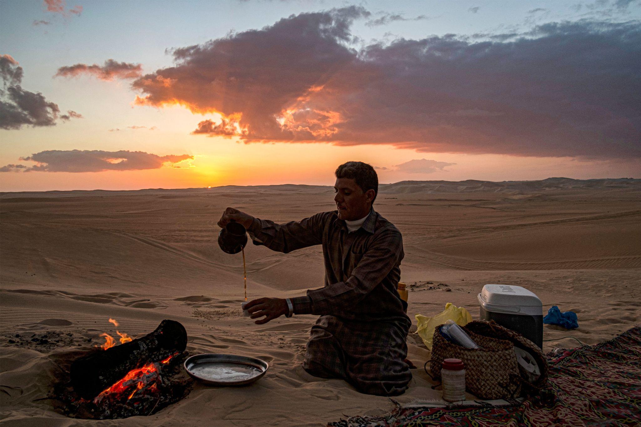 A man pours tea beside a campfire on Siwa's sand dunes at sunset