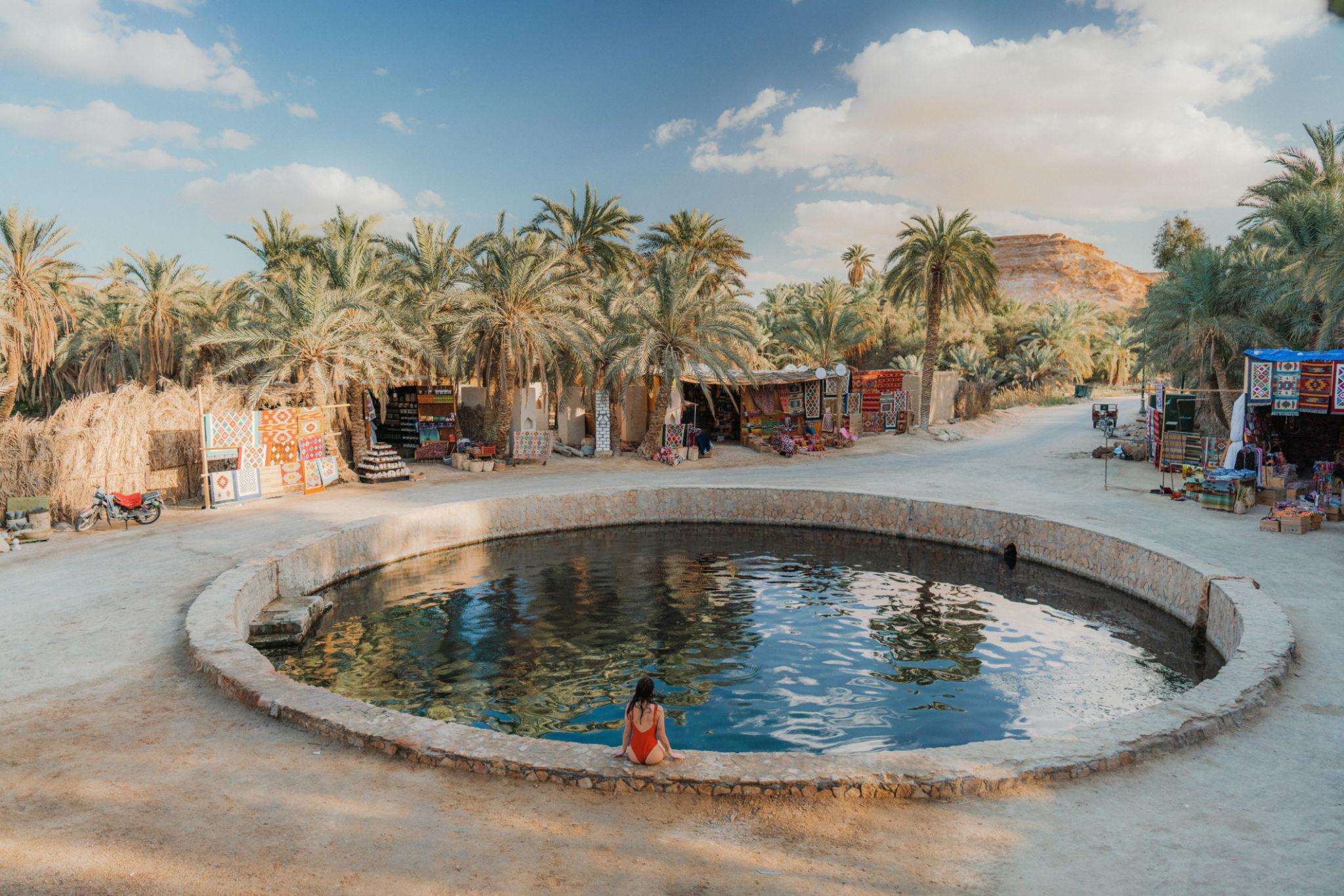 A woman in a red swimsuit sits at the edge of a round natural spring pool surrounded by palm trees and colorful market stalls
