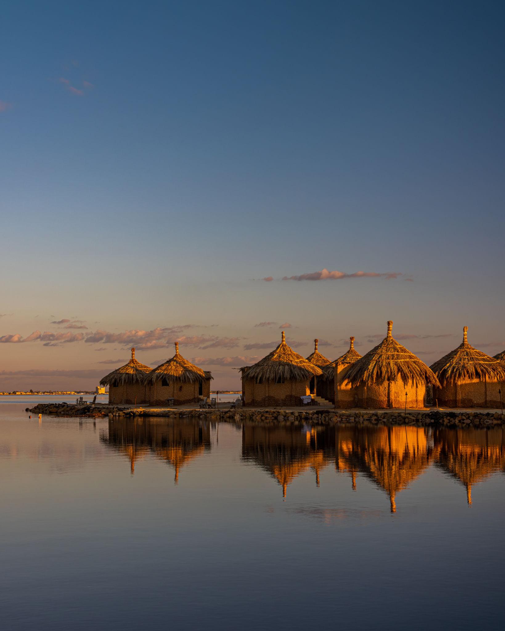 Thatched-roof huts reflected in a calm Siwa lake at sunset