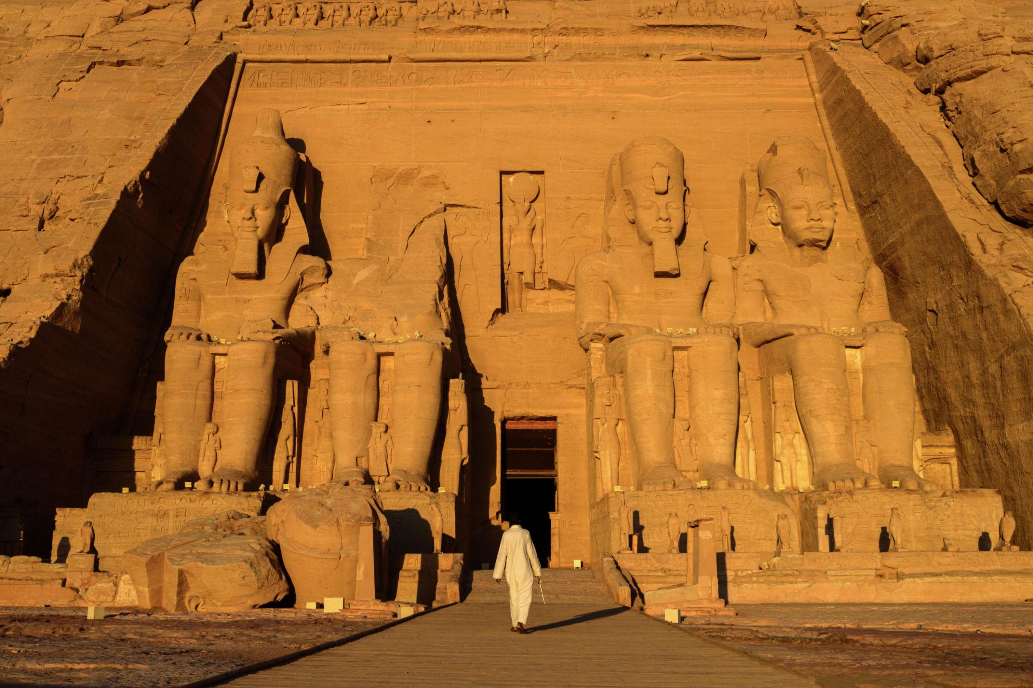 A lone figure in white walks toward the colossal sandstone statues of Abu Simbel at golden hour.