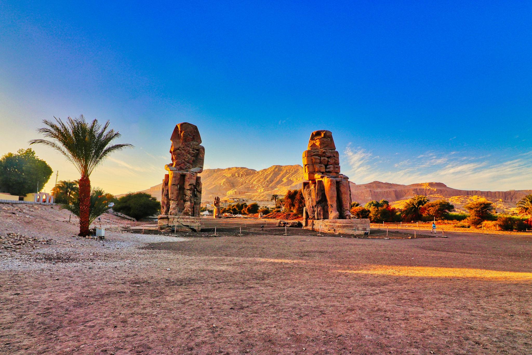 Colossi of Memnon framed by a palm tree against desert cliffs on Luxor's west bank