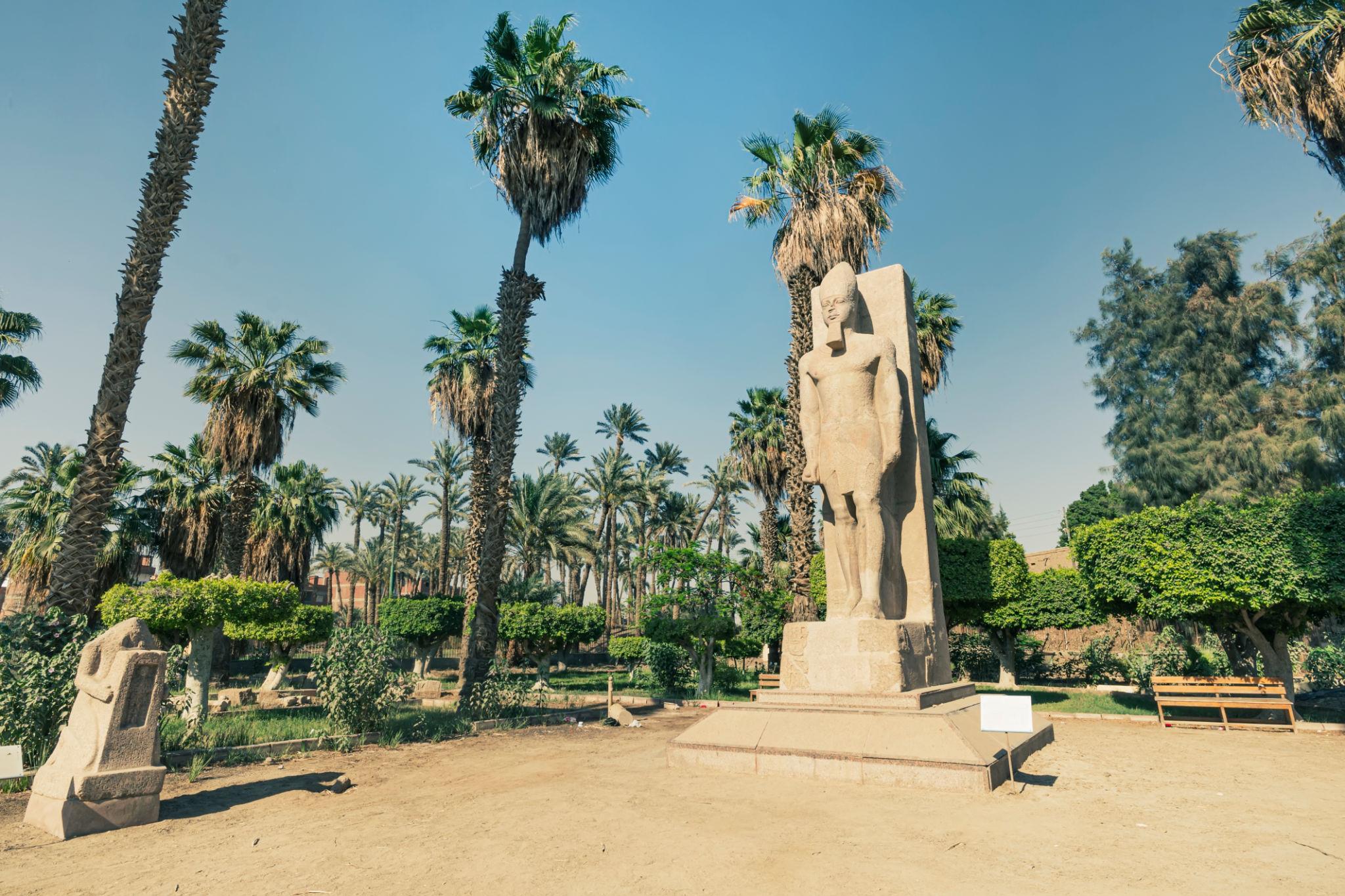 Ancient Egyptian standing pharaoh statue on a pedestal surrounded by palm trees and greenery