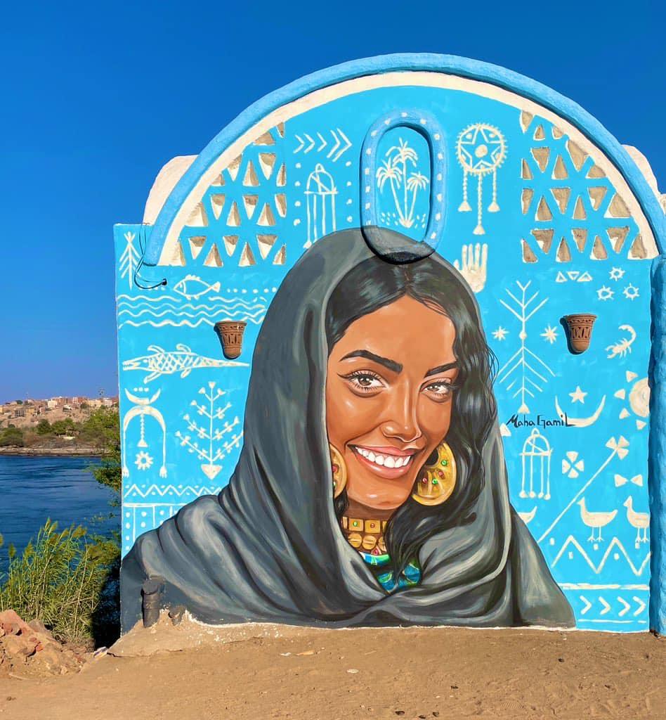 Vibrant blue mural of a smiling Nubian woman in a dark veil with gold jewelry by the Nile