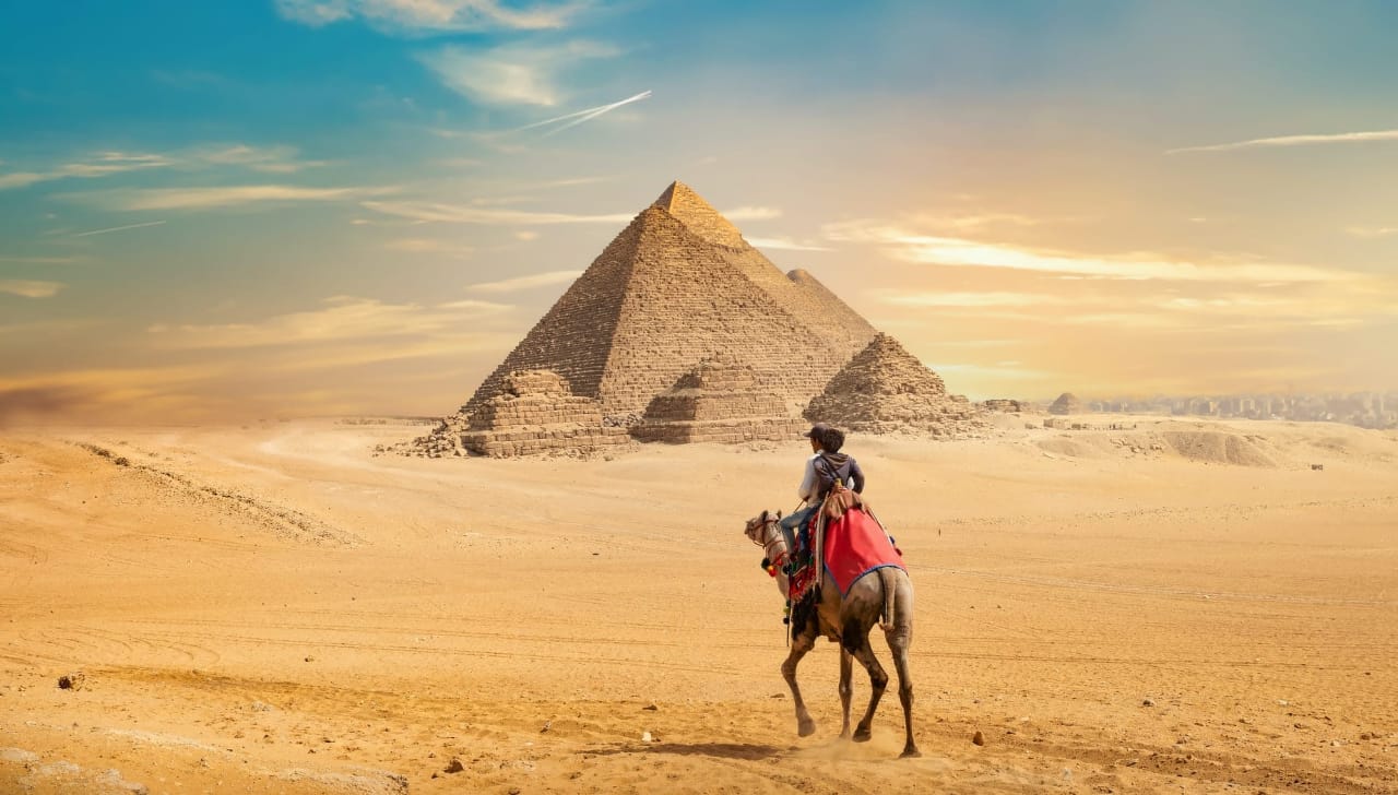 Lone camel rider crossing the desert toward the pyramids of Giza at sunset