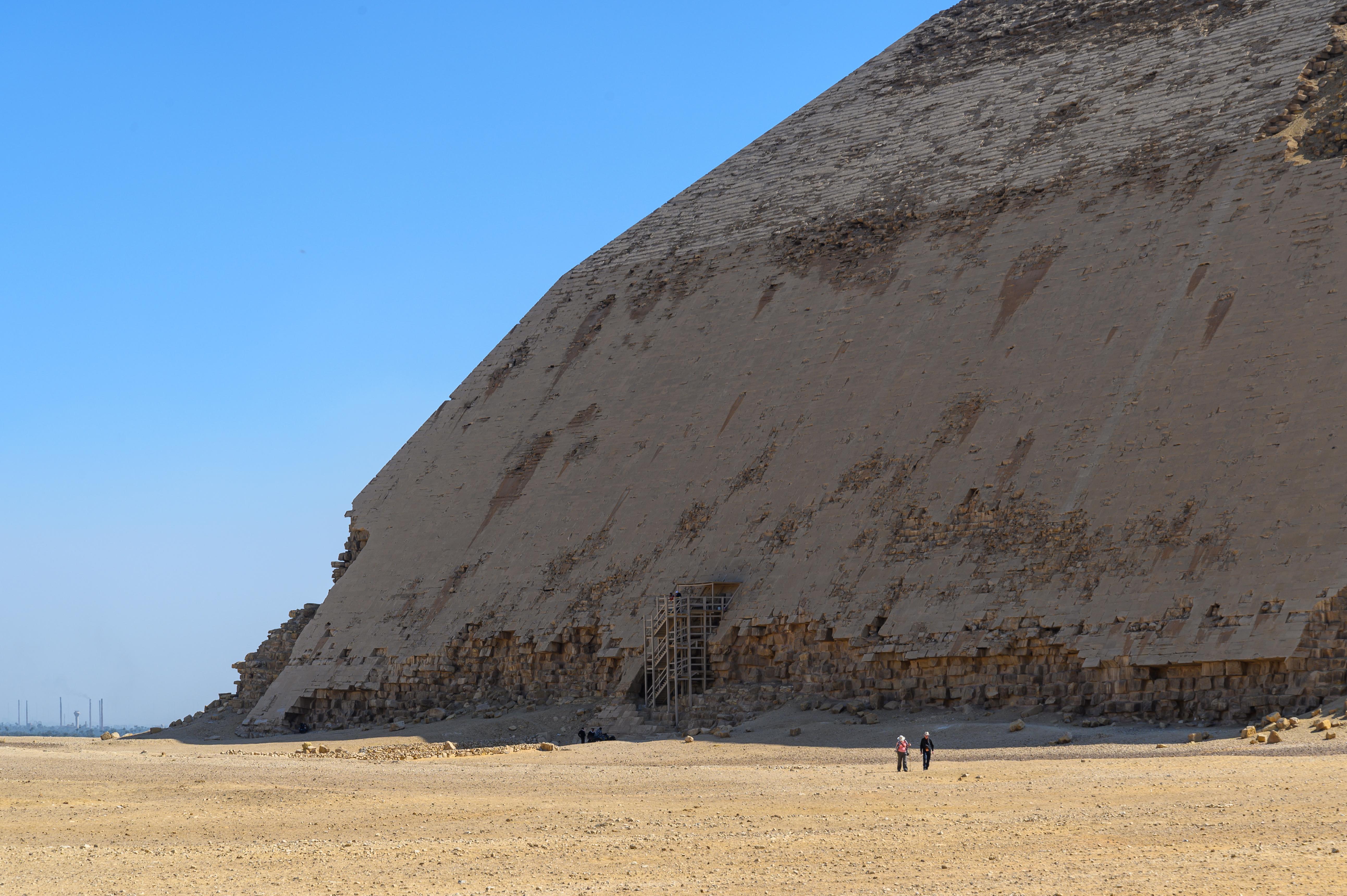 Two tiny visitors stand before the vast weathered stone face of the Bent Pyramid at Dahshur