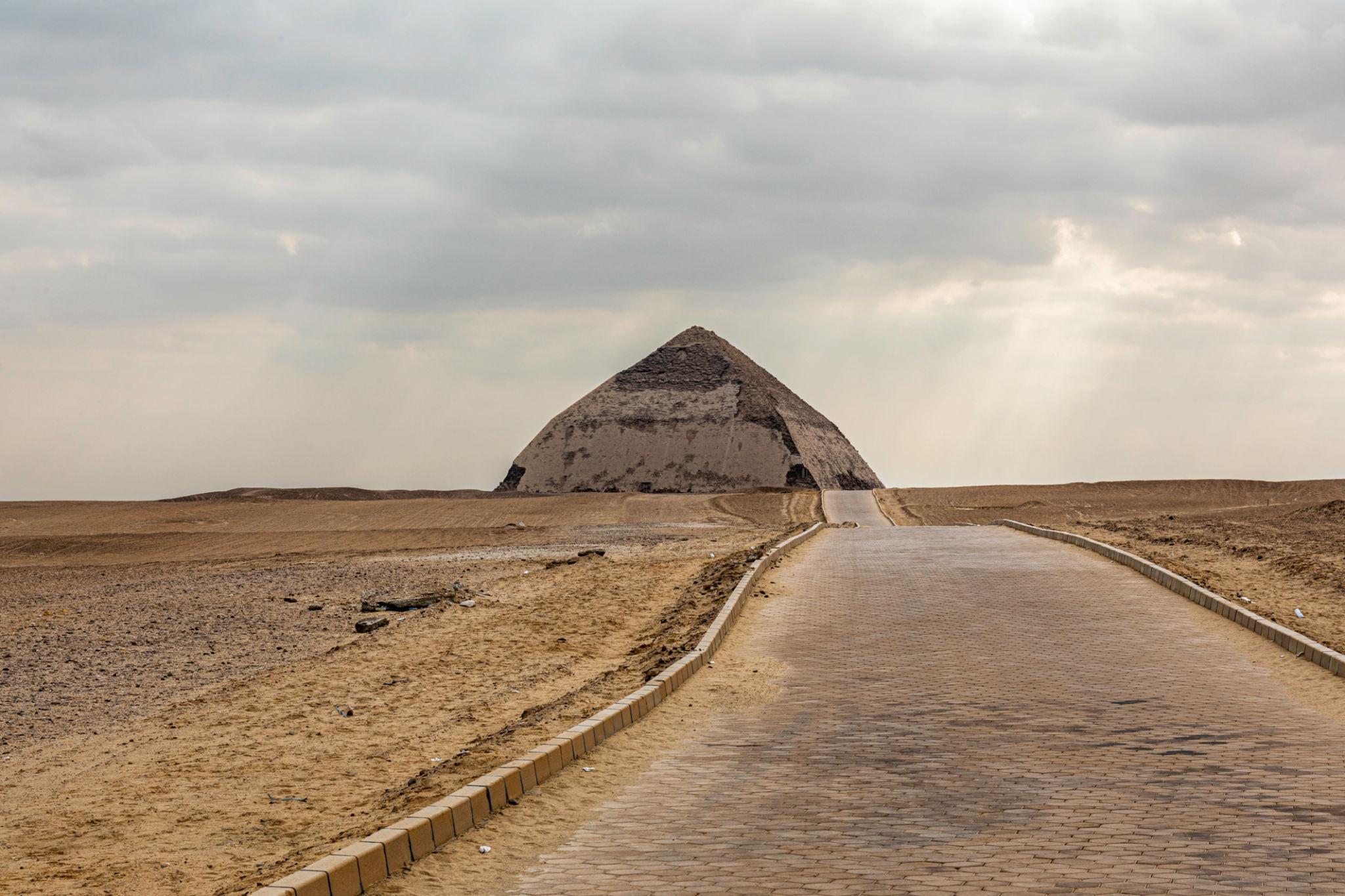 A brick causeway leads straight toward the Bent Pyramid rising from the flat desert plain.