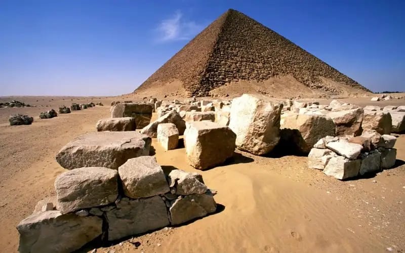 Large limestone blocks scattered in desert sand before the Red Pyramid of Dahshur under blue sky