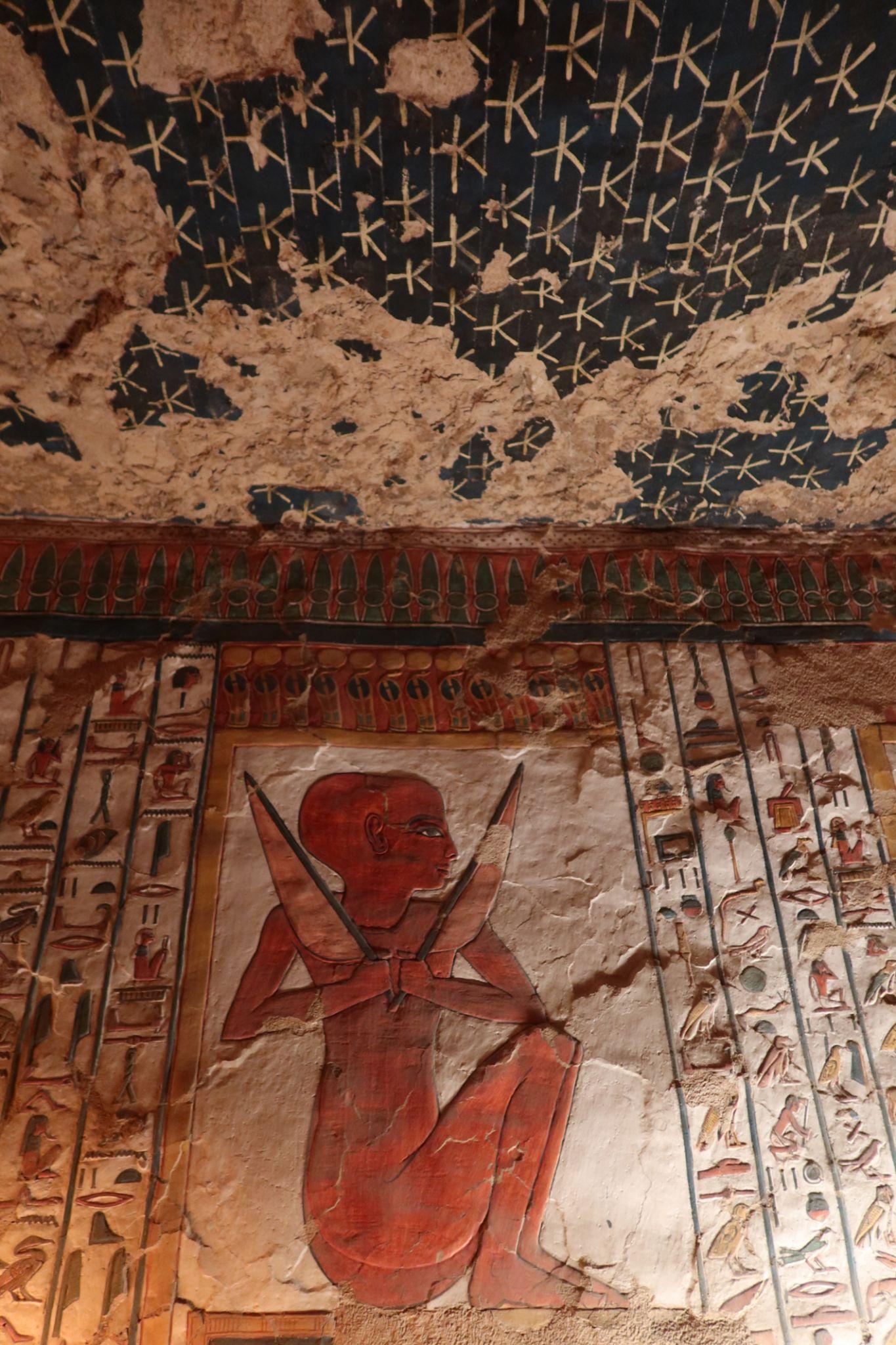 Red-painted mummiform deity on a tomb wall under a starred blue ceiling.