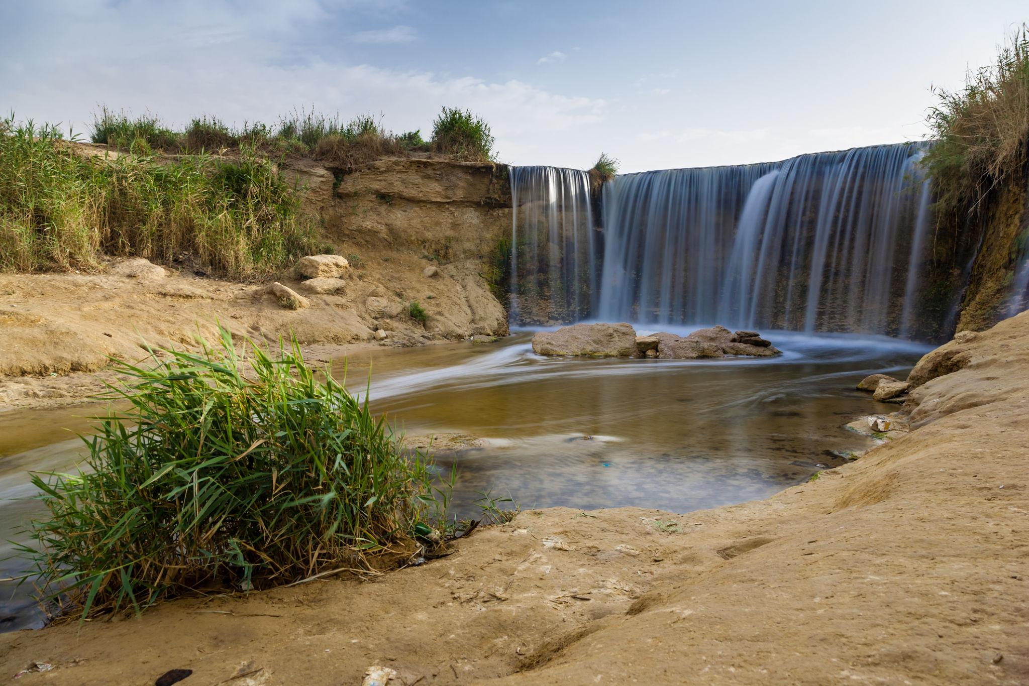 Long-exposure waterfall flowing past reeds and sandy desert banks